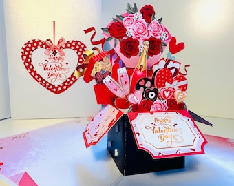 May include: A pop-up Valentine's Day card featuring a red and white polka-dot heart with the text "Happy Valentine's Day." The card includes a bouquet of roses, champagne, and heart-shaped gifts. The card is in a black box.