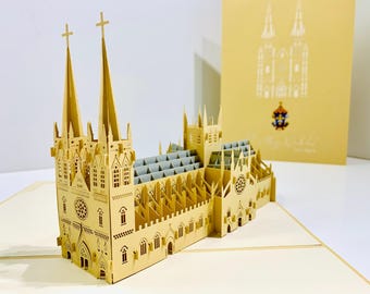 Puede incluir: Maqueta de papel cortada con láser de una catedral, con dos altas agujas rematadas con cruces, ventanas detalladas y un tejado gris. En el fondo, una tarjeta a juego con una ilustración de la catedral. El modelo está hecho de papel de color beige.
