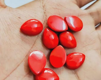 May include: A collection of glossy, teardrop-shaped, bright red beads. The beads are smooth and reflect light, suggesting a polished surface. They are held in a hand, showcasing their size and color.