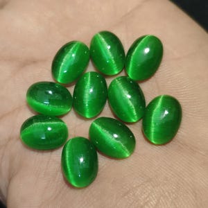 Op de afbeelding: Negen ovaalvormige cabochon edelstenen in een levendige smaragdgroene kleur. Elke steen heeft een duidelijk chatoyant kattenoogeffect, met een heldere, lichtgevende band die verticaal over het gladde, gepolijste en bolle oppervlak loopt.