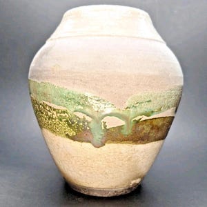 Könnte beinhalten: Eine handgefertigte Keramikvase mit einer einzigartigen, strukturierten Oberfläche. Die Vase hat einen cremefarbenen oberen Teil, der in einen hellbraunen Mittelteil mit grünen und braunen Akzenten übergeht. Der Sockel ist dunkelbraun.