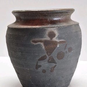 May include: A dark grey ceramic vase with a wide base and a narrow neck. The rim is a reddish-brown colour. A brown abstract design of a person playing a sport is on the front of the vase.