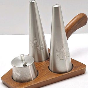 Puede incluir: Un juego de salero y pimentero vintage con azucarero, todo en metal plateado. Los saleros y el azucarero cónicos presentan diseños grabados y se asientan sobre una bandeja de madera con asa. Un juego de utensilios de cocina clásico.