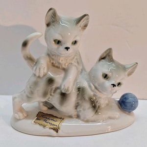 Może przedstawiać: Ceramiczna figurka przedstawiająca dwa bawiące się kocięta. Jedno kocię siedzi na drugim, z niebieską piłką w pobliżu. Kocięta są głównie białe z szarymi znaczeniami i zielonymi oczami. Podstawa jest biała, a etykieta głosi "Handarbeit aus Thüringen."