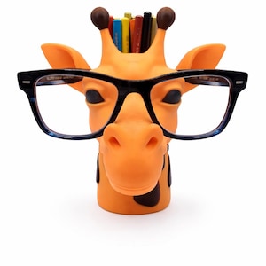 May include: Orange giraffe-shaped desk organizer with black-framed glasses. The giraffe has brown spots and holds pens and pencils. A fun and functional desk accessory for home or office.