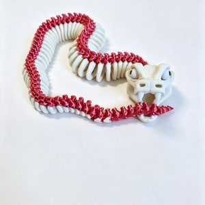 May include: An articulated snake figurine featuring a white head with prominent fangs and eyes, connected to a segmented body. The body consists of alternating white and metallic red plastic links that allow for flexible movement and positioning.