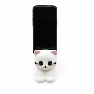Könnte beinhalten: Ein weißer, katzenförmiger Telefonständer mit rosa Akzenten, der ein schwarzes Smartphone hält. Der Ständer hat große schwarze Augen, rosa Innenohren und kleine rosa Rouge-Markierungen. Das Telefon steht aufrecht.