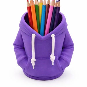 Peut inclure: Un pot à crayons violet en forme de sweat à capuche, avec des cordons blancs et une poche avant. Il est rempli de crayons de couleur, notamment rouge, orange, jaune, vert, bleu, rose et violet. Les crayons sont taillés.