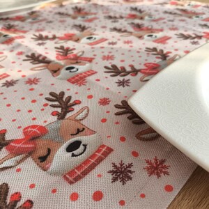 May include: A festive table runner featuring a repeating pattern of reindeer with red scarves and snowflakes on a light pink background. The runner is made of a textured fabric and is partially covered by a white plate.