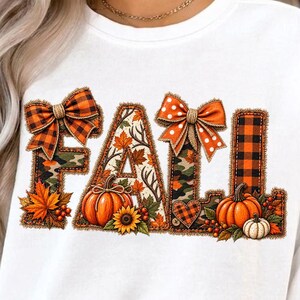 Op de afbeelding: Een wit sweatshirt met lange mouwen en het woord FALL in grote blokletters. De letters zijn versierd met oranje pompoenen, zonnebloemen, herfstbladeren, ruitpatronen, camouflageprints en twee oranje strikken met stippen.