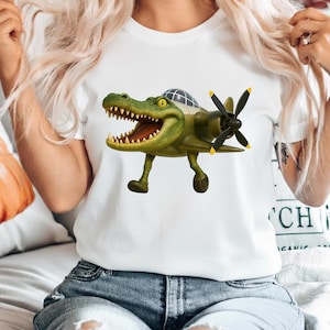 May include: A white t-shirt featuring a graphic of a green crocodile with an open mouth, designed with the body of a vintage propeller airplane. The creature has yellow eyes, sharp teeth, and a cockpit canopy on its back, with black propellers.