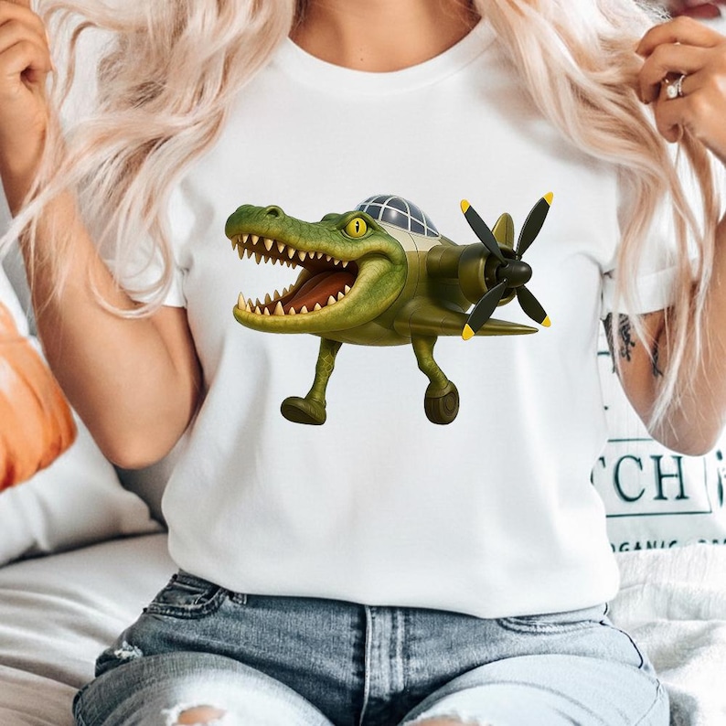May include: A white t-shirt featuring a graphic of a green crocodile with an open mouth, designed with the body of a vintage propeller airplane. The creature has yellow eyes, sharp teeth, and a cockpit canopy on its back, with black propellers.