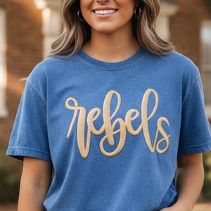 May include: A blue t-shirt with the word "rebels" in gold script. The shirt is short-sleeved and has a relaxed fit. The person is wearing blue jeans and a gold bracelet.