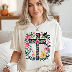 Könnte beinhalten: Cremefarbenes T-Shirt mit einem schwarzen Kreuzdesign, das mit einem floralen Kranz aus rosa, gelben und grünen Blumen verziert ist. Das Shirt hat eine lockere Passform und ist für den Freizeitgebrauch geeignet. Das Design ist mittig auf dem Shirt platziert.