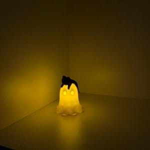 Könnte beinhalten: Ein leuchtendes, gelbes Nachtlicht in Geisterform mit schwarzen Haaren und leuchtend gelben Augen. Das Licht steht auf einer weißen Oberfläche in einer Ecke und strahlt warm.