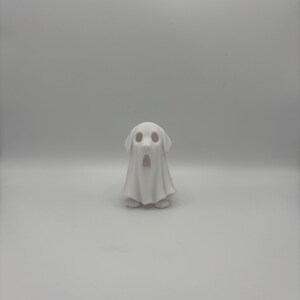 May include: A white ceramic figurine of a dog wearing a ghost costume. The dog's face is visible through the ghost's eye holes, and its paws are visible at the bottom. The ghost costume has a draped appearance.
