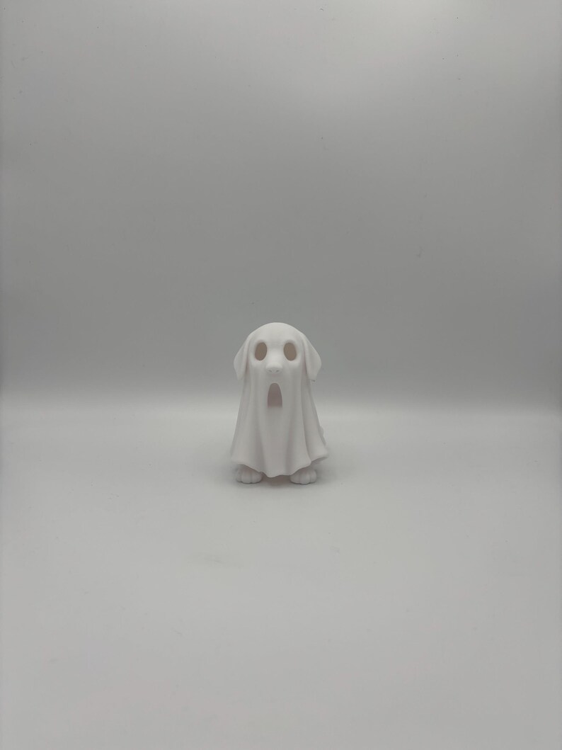 May include: A white ceramic figurine of a dog wearing a ghost costume. The dog's face is visible through the ghost's eye holes, and its paws are visible at the bottom. The ghost costume has a draped appearance.