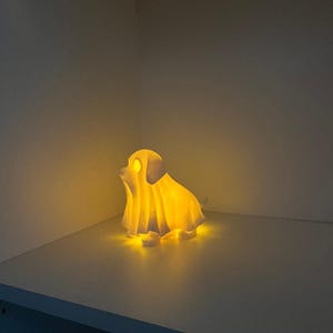 May include: A glowing, yellow-lit dog-shaped lamp, resembling a ghost. The lamp is on a white shelf, casting a warm glow in a corner. The dog has a draped sheet over it, with its ears and face visible.