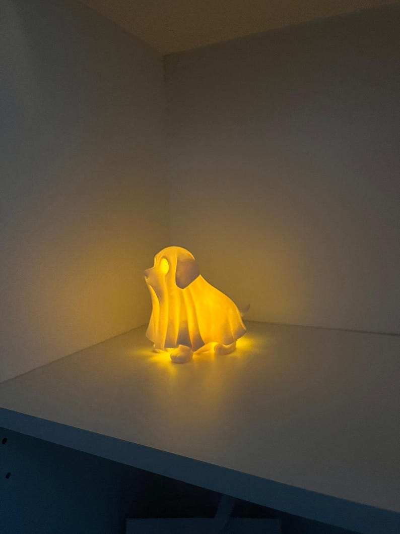 May include: A glowing, yellow-lit dog-shaped lamp, resembling a ghost. The lamp is on a white shelf, casting a warm glow in a corner. The dog has a draped sheet over it, with its ears and face visible.