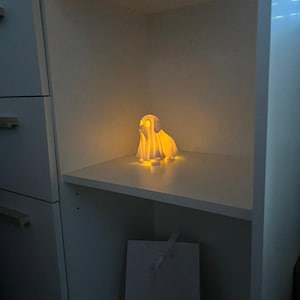 May include: A glowing, white, ghost-shaped lamp sits on a shelf inside a white cabinet. The lamp emits a warm, yellow light, illuminating the shelf. A white gift bag with a ribbon is below the shelf. The overall theme is home decor.