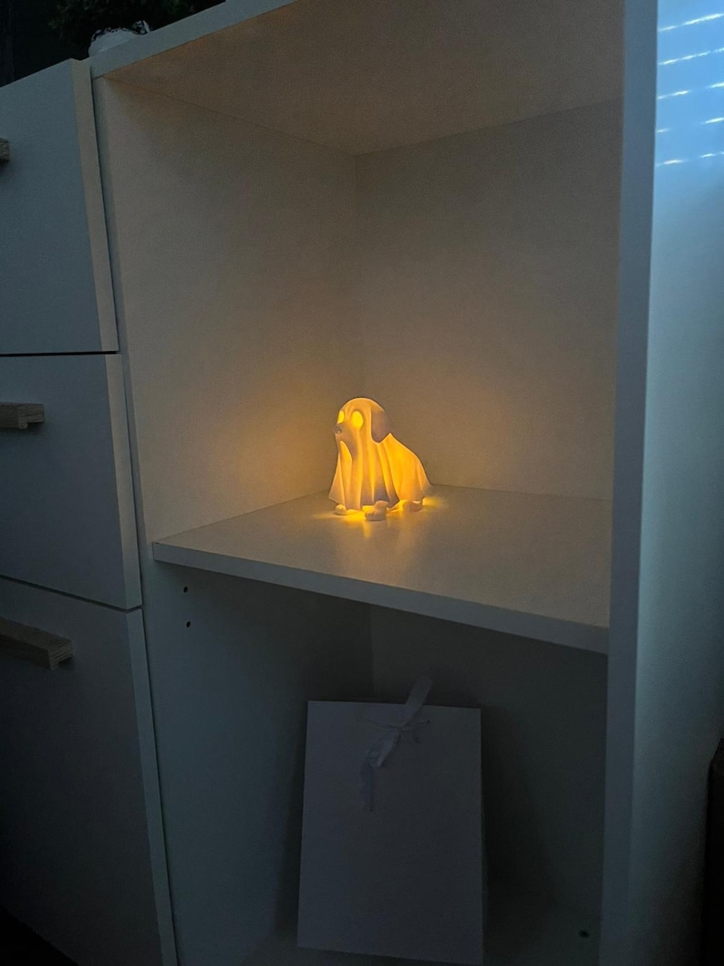 May include: A glowing, white, ghost-shaped lamp sits on a shelf inside a white cabinet. The lamp emits a warm, yellow light, illuminating the shelf. A white gift bag with a ribbon is below the shelf. The overall theme is home decor.