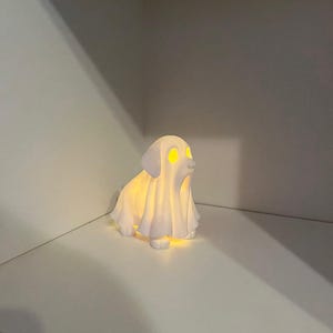 May include: A white ceramic dog-shaped night light with glowing yellow eyes and a draped sheet-like form. The light emits a soft glow, creating a whimsical and spooky ambiance. Perfect for Halloween or a dog lover's decor.