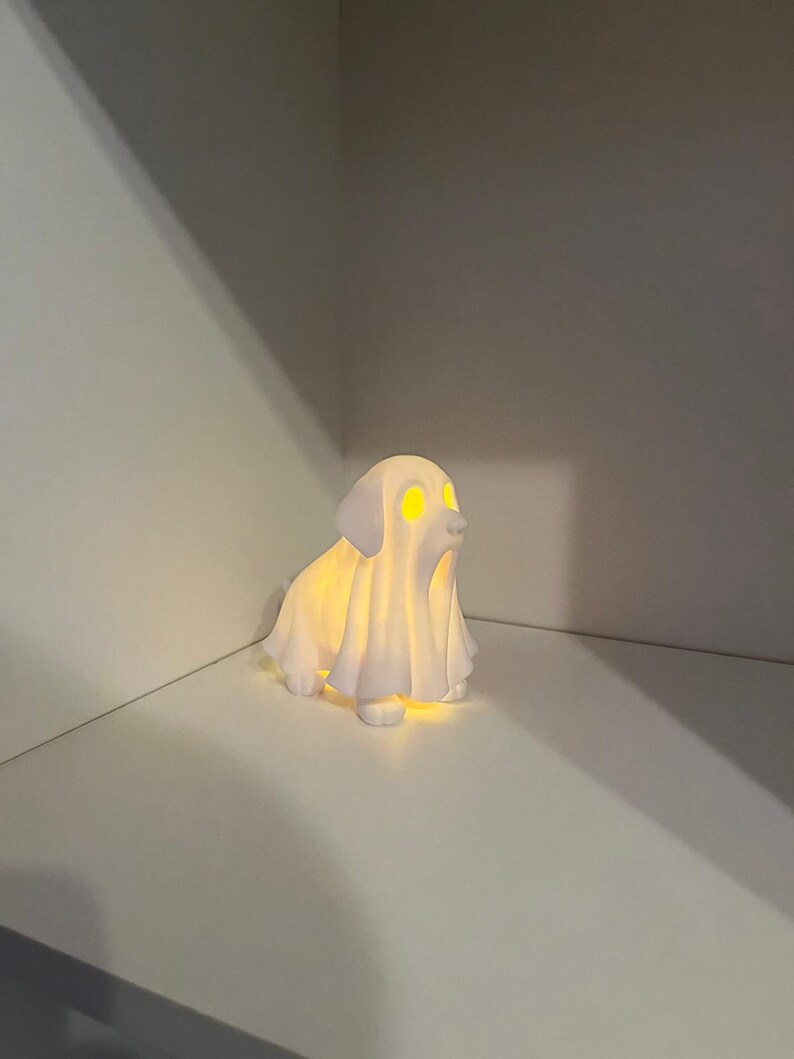 May include: A white ceramic dog-shaped night light with glowing yellow eyes and a draped sheet-like form. The light emits a soft glow, creating a whimsical and spooky ambiance. Perfect for Halloween or a dog lover's decor.