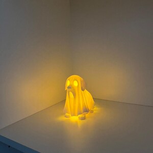 May include: A glowing, white dog-shaped lamp with a ghost-like appearance. The lamp emits a warm, yellow light, illuminating the dog's eyes and the surrounding area. The lamp is placed on a white surface.