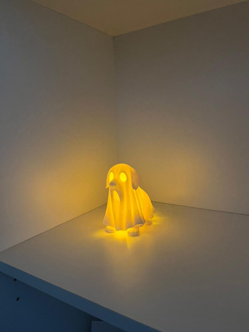 May include: A glowing, white dog-shaped lamp with a ghost-like appearance. The lamp emits a warm, yellow light, illuminating the dog's eyes and the surrounding area. The lamp is placed on a white surface.