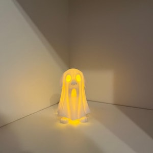 May include: A white ghost-shaped lamp with glowing yellow light emanating from within. The lamp has a draped sheet design with visible eyes and a mouth. The lamp is sitting on a white surface.