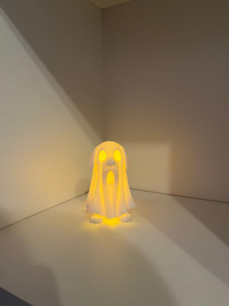 May include: A white ghost-shaped lamp with glowing yellow light emanating from within. The lamp has a draped sheet design with visible eyes and a mouth. The lamp is sitting on a white surface.