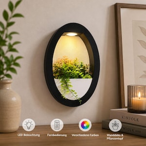 Può includere: Una fioriera da parete ovale con cornice nera e bacino bianco contenente fogliame artificiale verde. L'interno presenta un pannello posteriore strutturato illuminato da una calda luce LED. Quattro icone circolari in basso mostrano il testo: LED Beleuchtung, Fernbedienung, Verschiedene Farben e Wanddeko & Pflanzentopf.