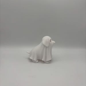 May include: A small, white figurine of a dog covered in a sheet, resembling a ghost. The dog is in a sitting position with its head turned to the side. The figurine is made of a smooth, solid material.