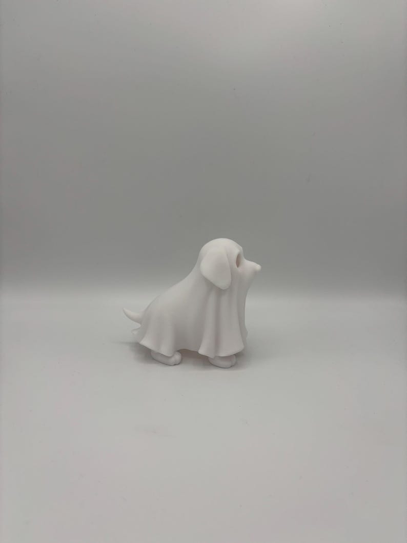 May include: A small, white figurine of a dog covered in a sheet, resembling a ghost. The dog is in a sitting position with its head turned to the side. The figurine is made of a smooth, solid material.