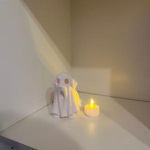 May include: A white ceramic dog figurine draped with a ghost sheet, next to a lit candle. The dog has a long snout and floppy ears. The candle is in a white holder and emits a warm glow, creating a spooky Halloween theme.