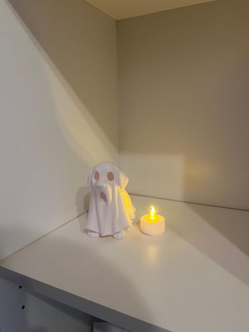 May include: A white ceramic dog figurine draped with a ghost sheet, next to a lit candle. The dog has a long snout and floppy ears. The candle is in a white holder and emits a warm glow, creating a spooky Halloween theme.