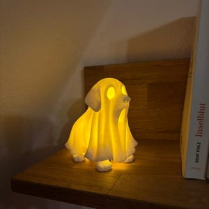 May include: A glowing white dog-shaped lamp with a draped design, emitting a warm yellow light. The lamp is placed on a wooden shelf, creating a cozy ambiance. The dog's eyes and nose are illuminated, adding a whimsical touch.