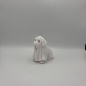 May include: A white ceramic figurine of a dog wearing a ghost costume. The dog is sitting with its mouth open, and the ghost costume drapes over its body. The figurine is on a white background.