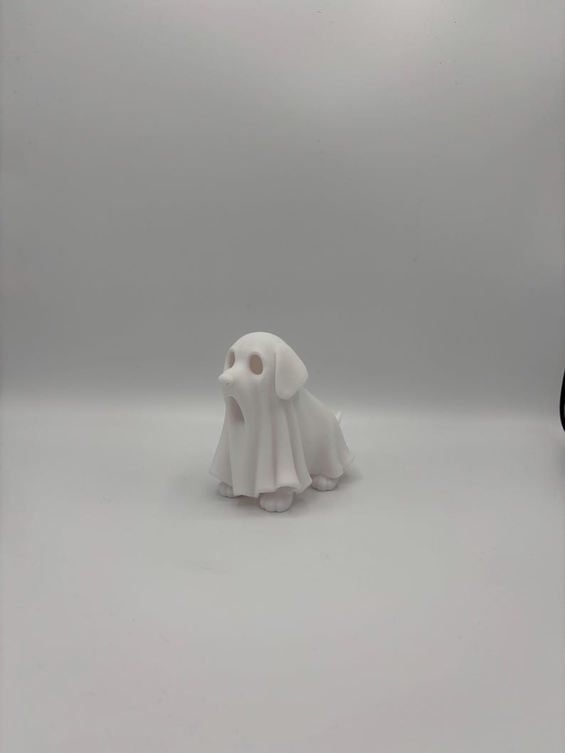 May include: A white ceramic figurine of a dog wearing a ghost costume. The dog is sitting with its mouth open, and the ghost costume drapes over its body. The figurine is on a white background.