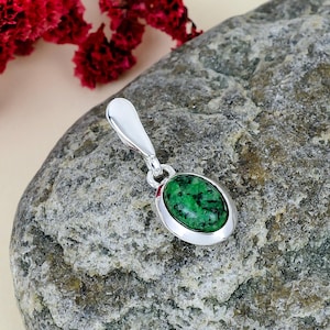 Op de afbeelding: Een zilveren hanger met een ovaalvormige groene steen met zwarte insluitingen. De hanger heeft een gepolijst zilveren oogje aan de bovenkant, ontworpen om een ketting doorheen te halen. De steen is gezet in een gladde, afgeronde zilveren rand.