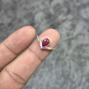 May include: A silver V-shaped ring featuring a single pear-cut, deep red gemstone set in a bezel. The band has a polished metallic finish and follows a chevron silhouette, centering the faceted red stone at the point of the V-shaped design.