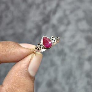 Può includere: Un anello in argento con una pietra preziosa centrale a forma di pera, sfaccettata e di colore rosa intenso. La fascia in metallo presenta intricati dettagli decorativi a volute su entrambi i lati della montatura, creando un contrasto cromatico.