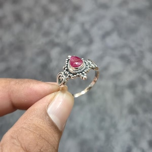 May include: A silver ring featuring a central teardrop-shaped faceted pink gemstone. The metal band displays intricate oxidized detailing, including small beaded accents and a heart-shaped motif on the side, creating a textured appearance.