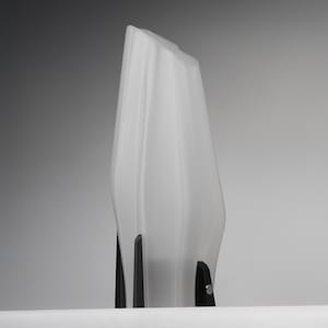 May include: A tall, white, sculptural vase with a textured surface. The vase has a unique, elongated shape and is supported by a black base with three vertical supports. The design is modern and minimalist.