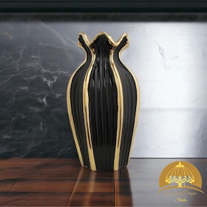 Puede incluir: Un jarrón de cerámica negro y dorado con un diseño acanalado. El jarrón tiene un acabado brillante con rayas doradas verticales y un borde festoneado. Es un elemento decorativo para el hogar.