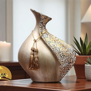 May include: A decorative vase with a gold and silver floral design. The vase has a unique shape with a wide base and a narrow neck. It is adorned with gold tassels and is approximately 12 inches tall.