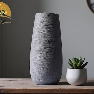 Op de afbeelding: Een hoge, grijze vaas met textuur en een golvende rand, naast een kleine witte pot met een groene vetplant. De vaas heeft een ruw, geribbeld oppervlak. De items staan op een houten tafel.