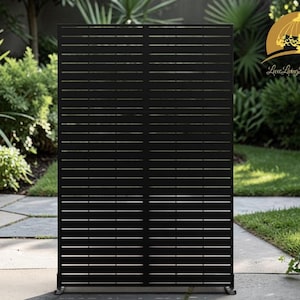 May include: A black outdoor privacy screen with horizontal slats. The screen is made of a dark material and is set against a backdrop of greenery and a paved patio. The design provides both privacy and a decorative element to the outdoor space.