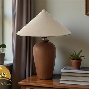 May include: A brown ceramic table lamp with a cream-colored lampshade. The lamp has a rounded base and a conical shade. The lamp is on a wooden table, next to a window and a small potted plant. The lamp is approximately 24 inches tall.