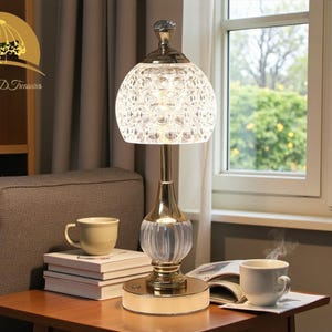 May include: A decorative table lamp with a gold base and a clear, patterned glass shade. The lamp is turned on, illuminating the room. A cup of coffee and books are on the table. The lamp is from Luxe Living D Treasures.